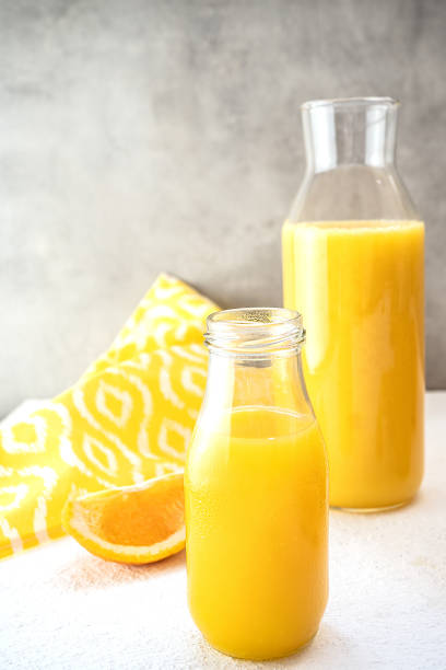 ガラス瓶と搾りたてのオレンジジュースの入ったデカンタが、灰色のコンクリートの壁の白い面に立っています。近くにはオレンジのスライスと白い模様の黄色いタオルが1枚あります。垂直 - juice carafe glass decanter ストックフォトと画像