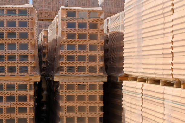 Stack of red bricks arranged on wooden pallet standing inside unfinished building under construction, exposed brick walls and concrete floor visible in background, no people present