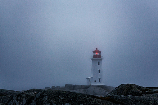 Peggy's Cove Lighthouse is an iconic international landmark travel and tourist destination, located on the extreme terrain rocky coastline of the maritime province of Nova Scotia, Canada, on the Atlantic Ocean. Here, the lighthouse warning beam is trying to penetrate through very thick fog on a mid-October autumn night just after evening sunset.