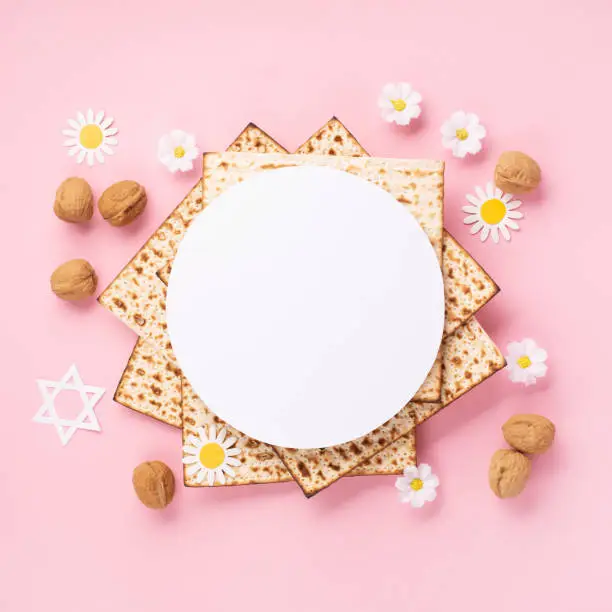 Jewish holiday Passover greeting card concept with matzah, nuts, tulip and daisy flowers on pink table. Seder Pesach spring holiday background, top view, copy space.