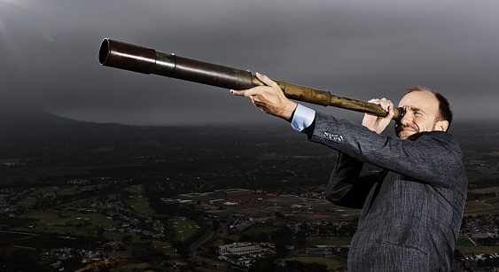 Man wearing a suit looks into the distance through a brass nautical telescope.