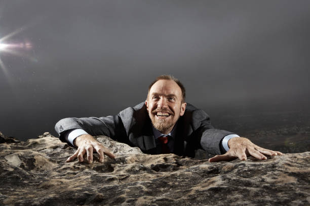 Success! Well-dressed businessman, climbing a mountain, is happy to have reached the peak Smiling businessman in a suit climbs to the top of a mountain, representing achievement and accomplishment. reach destination stock pictures, royalty-free photos & images