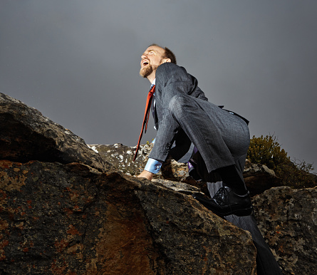 Businessman wearing a suit clambers to the top of a rocky mountain, representing triumph and accomplishment.