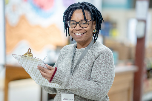A senior woman of African decent stands in the middle of a warehouse with a clipboard in hand as she fills out some paper work.  She is dressed casually and has a name badge around her neck as she works away volunteering her time at a local Food Bank.