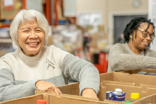 Two senior ladies work together as they volunteer their time at a local Food Bank.  They are both dressed casually and smiling as they pack the cardboard boxes on the table in front of them with non-perishable food items.  Both are smiling as they happily give of their time for the cause.