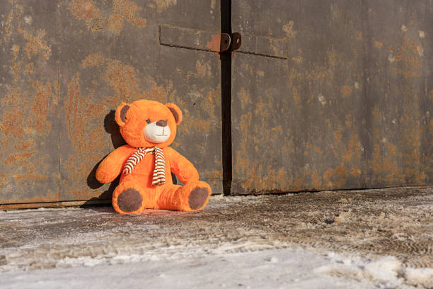 Lost orange teddy bear toy sitting on muddy sidewalk against rusty gate Lost orange teddy bear toy sitting on muddy sidewalk covered with dirty snow against rusty gate. Loneliness and sadness concept. behavior teddy bear doll old stock pictures, royalty-free photos & images