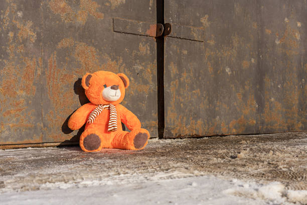 zagubiona pomarańczowa zabawka z misiem siedząca na błotnistym chodniku przy zardzewiałej bramie - behavior teddy bear doll old zdjęcia i obrazy z banku zdjęć