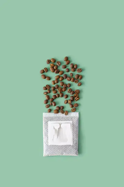 Photo of Drip coffee bag with ground coffee for brewing in cup and scattered coffee beans, pack with paper bag drip coffee filter, mint color background, top view, minimal flat lay with copy space