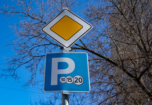 The main road sign and the parking sign on the same pole against the background of bare branches and blue sky.