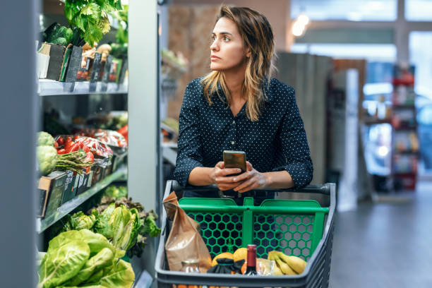 istock Modern woman using smartphone with trolley cart while walking and taking products from shelf in a grocery store. 1387514413