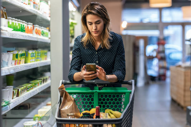 istock Modern woman using smartphone with trolley cart while walking and taking products from shelf in a grocery store. 1387514519