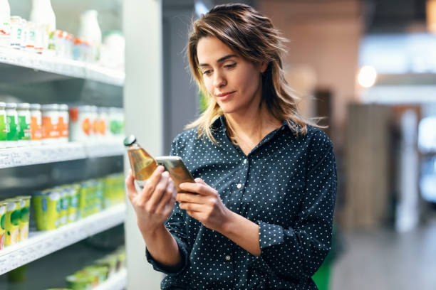 istock Modern woman using smartphone with trolley cart while walking and taking products from shelf at the grocery 1387515200