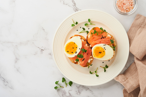 Sandwich with delicious toppings, smoked salmon, eggs, herbs and microgreens radish, black sesame seeds over white plate on white marble table background. Healthy open sandwich superfood. Top view.