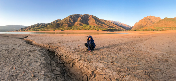 Desert. A teenager squatting on the cracked earth. Dried-up riverbed. Background of mountain.