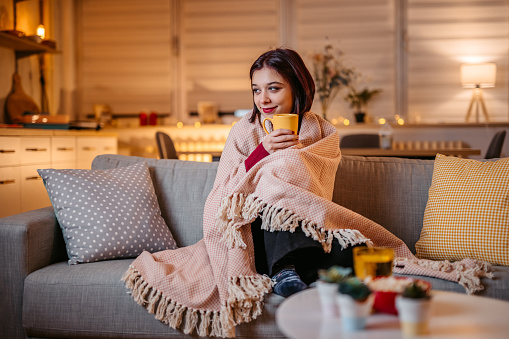 Beautiful young woman drinking hot tea wrapped in blanket on sofa at home.