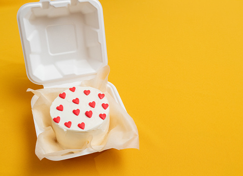 Bento cake in the form of circle with small red hearts in eco packaging. Picture for websites about desserts, food, confectionery. Yellow background with space for text. Selective focus.