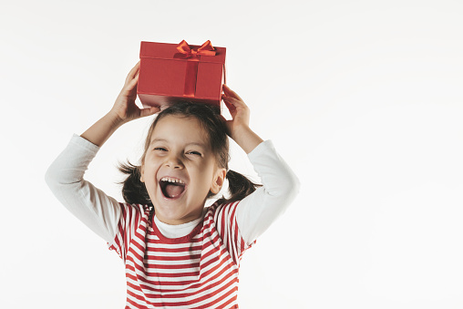 Caucasian girl is holding a red gift box in her hands over her head and is looking at camera with a toothy smile.