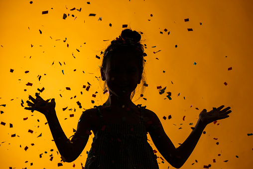 Silhouette of caucasian girl is standing under confetti falling down in front of yellow background.