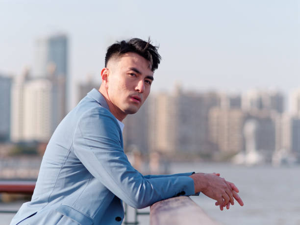 Portrait of handsome Chinese young man in light blue suit looking at camera with city buildings background, side view of confident businessman. stock photo