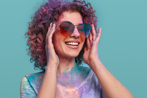 Self assured young female with curly hair and colorful powdered paints on body smiling and adjusting sunglasses against blue background during Holi festival