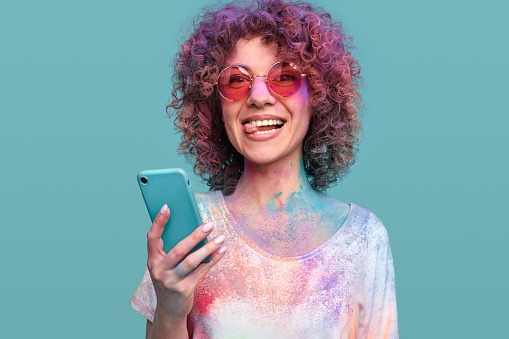 Funny millennial hipster female in sunglasses with colorful Holi paints on hair and skin making face and show tongue, while using mobile phone on blue background