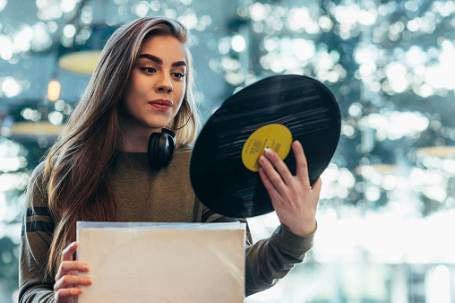 Beautiful woman holding a vinyl while selecting records in a shop. Young audiophile hipster woman in a record store. Vintage and retro style.