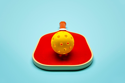 This is a photograph taken in the studio of a red pickleball paddle and yellow ball on a light blue background