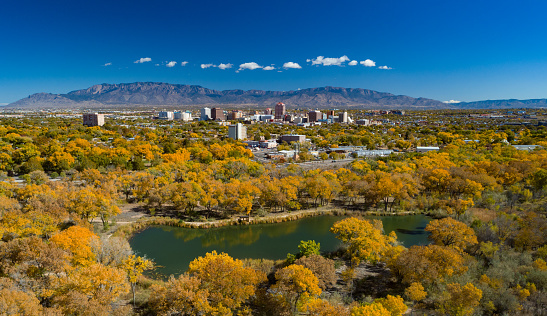 Albuquerque during autumn with the downtown skyline and the Sandia Mountains in the distance, with golden trees and a small lake in the foreground.