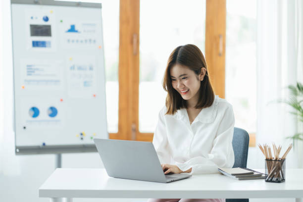 istock Smiling Asian female employee using laptop at workplace, looking at screen, focused businesswoman preparing economic report, working online project, cheerful intern doing computer work, typing. The graph on the whiteboard behind. 1387573038