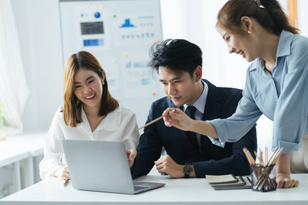 istock Cheerful asian people colleagues working together sit at desk look at computer screen discuss new project search solutions joking to increase effective communication concept, chart on whiteboard. 1387573699