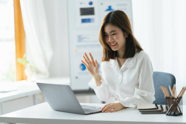 istock Excited asian lady having video call at work place, using laptop and graph on wihteboard behind, copy space. Happy woman waving at laptop screen, attending video conference. 1387576603