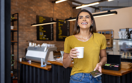 Happy Brazilian woman leaving a cafe after buying a cup of coffee to go and smiling - lifestyle concepts