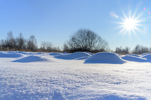 Deep, grainy snow that forms an uneven relief surface with the help of the wind. Close-up of snow, bottom view. Rainbow sun on the blue sky. In the distance, a rounded tree. Unusual shooting angle