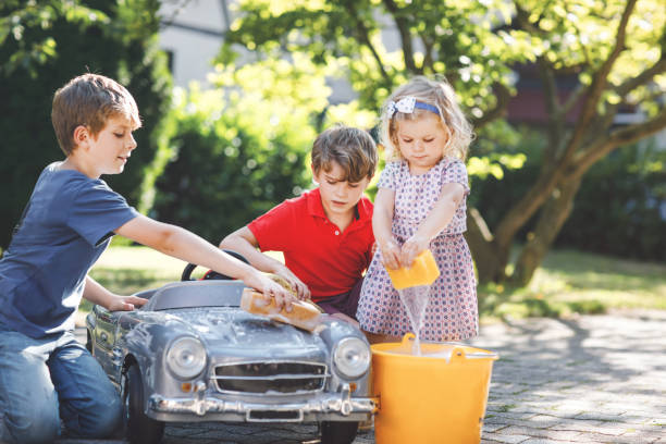 três crianças felizes lavando um carro de brinquedo grande e velho no jardim de verão, ao ar livre. dois meninos e uma garotinha limpando o carro com água e sabão, se divertindo com espirrar e brincar com esponja. - washing water car cleaning - fotografias e filmes do acervo