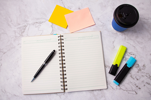 Top view of black pen on open book, blank notebook page, Yellow and blue highlighter pen, sticky note and blue cup of coffee on white marble table. marble office desk. template. Background.