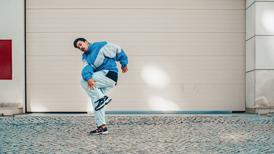 Active and Cheerful Young Adult Latin Man in Stylish Clothes Actively Dancing Hip Hop in Front of a Big Garage Door in an Urban Setting in a City. Scene Shot in a Small Town.