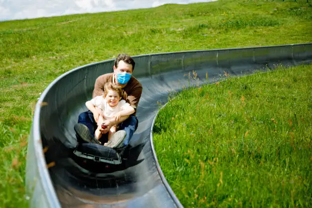 Little toddler girl and father having fun riding summer toboggan run sled down a hill. Active preschool child and dad with medical mask making funny activity otudoors. Family leisure with kids