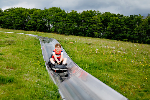 Young school kid boy having fun riding summer toboggan run sled down a hill in Hoherodskopf, Germany. Active child with medical mask making funny activity otudoors. Family leisure with kids.