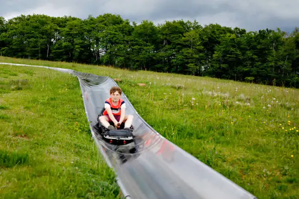 Young school kid boy having fun riding summer toboggan run sled down a hill in Hoherodskopf, Germany. Active child with medical mask making funny activity otudoors. Family leisure with kids