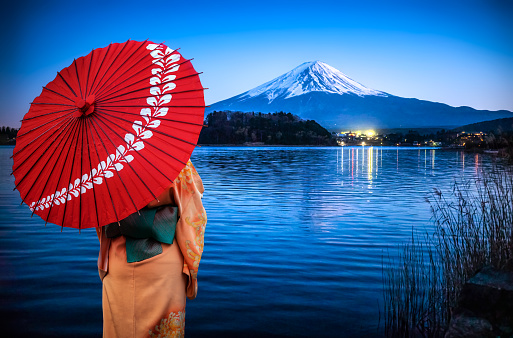 Rear view of a woman with traditional clothing with a red umbrella enjoying the view of Mount fuji at night reflected on Lake Kawaguchi, Japan