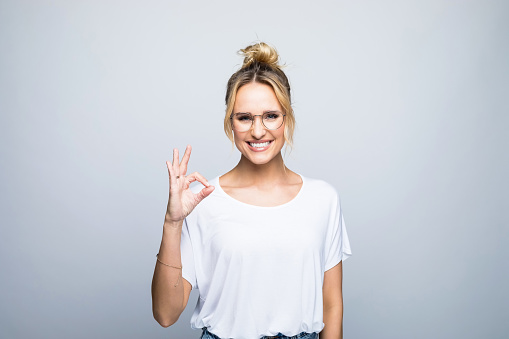 Portrait of happy beautiful blond woman showing OK sign and laughing at camera. Studio shot against gray background.