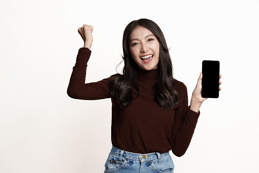 Portrait of beautiful young Asian woman holding smart phone, looking at camera and gasping with surprise expression on white background. Asian woman studio portrait advertising banner concept.