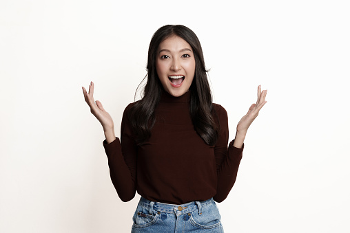 Portrait of beautiful young Asian woman showing expression of surprise on white background. Excited girl looking at camera and gasping with hands up beside.