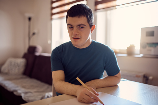 Young student with down syndrome drawing on paper at home and looking at camera.