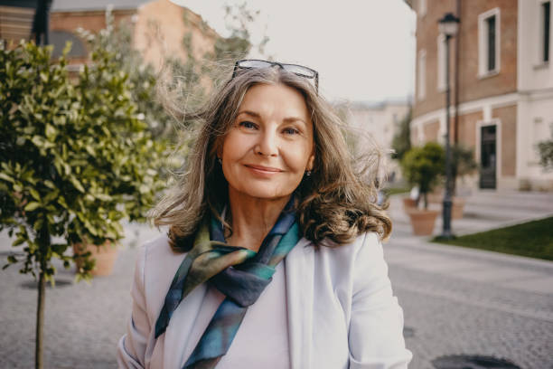 imagen al aire libre de una hermosa dama positiva con una sonrisa encantadora y cabello gris suelto disfrutando de un agradable día de verano, con chaqueta blanca, bufanda de seda y anteojos. belleza, estilo urbano y concepto de moda - mujeres mayores fotografías e imágenes de stock