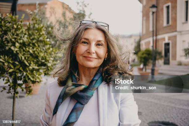 Outdoor Image Of Gorgeous Positive Lady With Charming Smile And Loose Gray Hair Enjoying Nice Summer Day Wearing White Jacket Silk Scarf And Eyeglasses Beauty Urban Style And Fashion Concept Stock Photo - Download Image Now