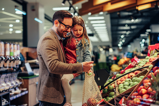 Smiling young adult man and his daughter choosing fruits and vegetables, putting it in canvas bag