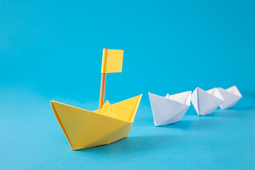 Red colored paper boat with flag is on blue background among other paper boats