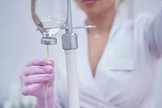 Cropped photo of doctor inserting needle through rubber gasket in inverted infusion glass bottle