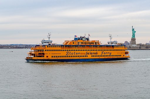 Yellow Staten Island Ferry in front Statue of Liberty National Park, New York City, during winter evening with overcast, horizontal